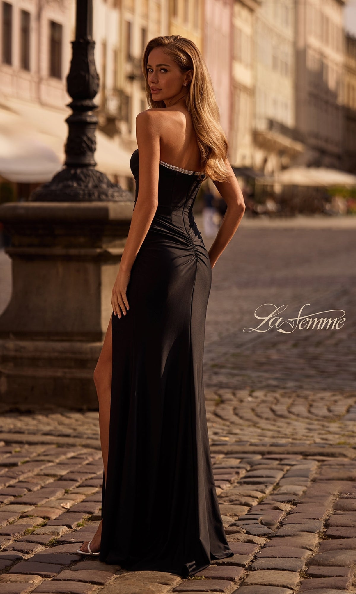 A woman wearing the Bead-Trimmed Strapless Prom Gown: La Femme 32756 poses on a cobblestone street, showing off the elegant thigh-high slit and sparkling bead details.